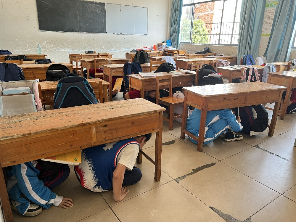 1747044429169654.jpg 学生们在老师的指挥下迅速抱头、闭眼,躲在各自的课桌下或墙角,尽量蜷曲身体,用书本等物品保护好头部.jpg